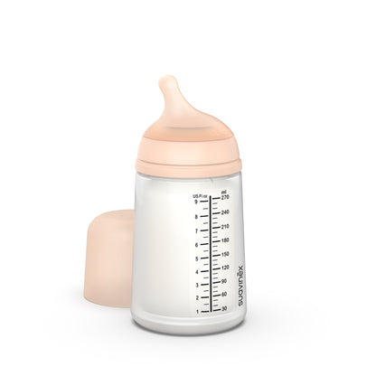 El Biberón Anticólicos ZERO ZERO 270ml Flujo Medio (M) de Suavinex, un biberón transparente con tapa rosa pálido y tetina de silicona, marcado en mililitros y onzas, se mantiene en posición vertical. La tapa, quitada, se encuentra a su lado: una pieza ideal para productos para bebés con entrega gratis en Mallorca.