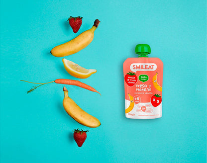 Una foto plana que muestra un Smileat - Pouch Fresa y Plátano Ecológico sobre un fondo azul, rodeado de fresas, plátanos, una rodaja de limón y una zanahoria, ideal para padres  que buscan productos aptos para bebés con opciones de entrega convenientes.